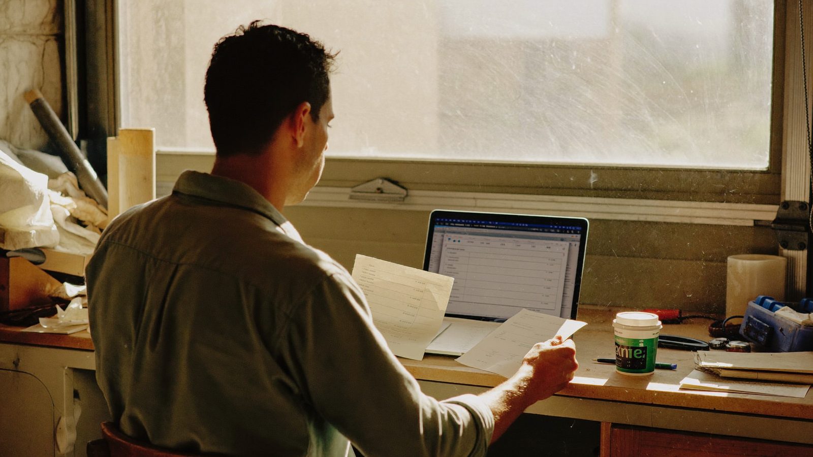 A small business owner reviews paper invoices and a laptop at a cluttered workshop desk in morning light