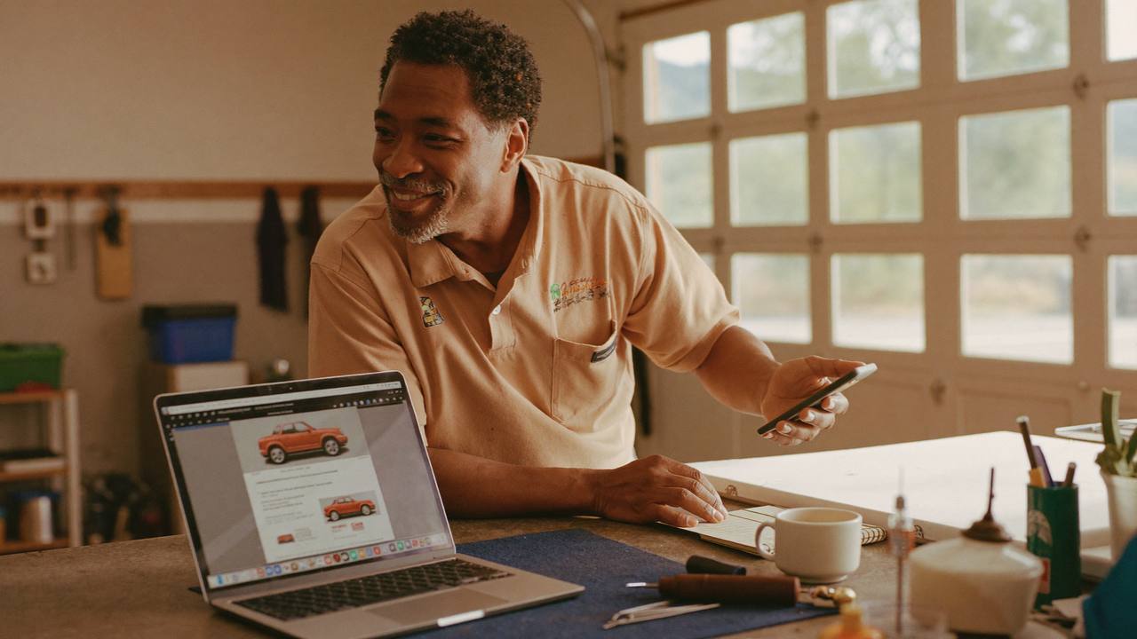 A small business owner checking a laptop and a phone side by side at a bright counter, coordinating marketing across more than one channel