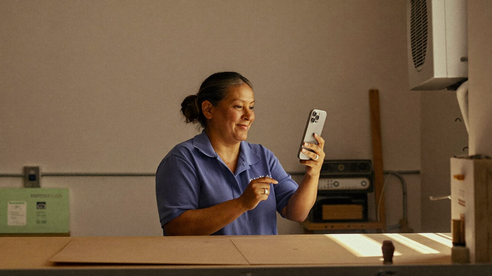 A small business owner smiles while reading a customer review on a smartphone at a plain work counter in warm morning light