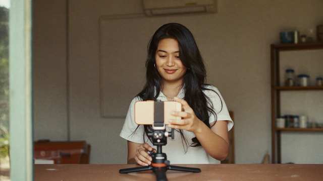 A small business owner films a short vertical video on a phone propped on a tripod stand inside their shop