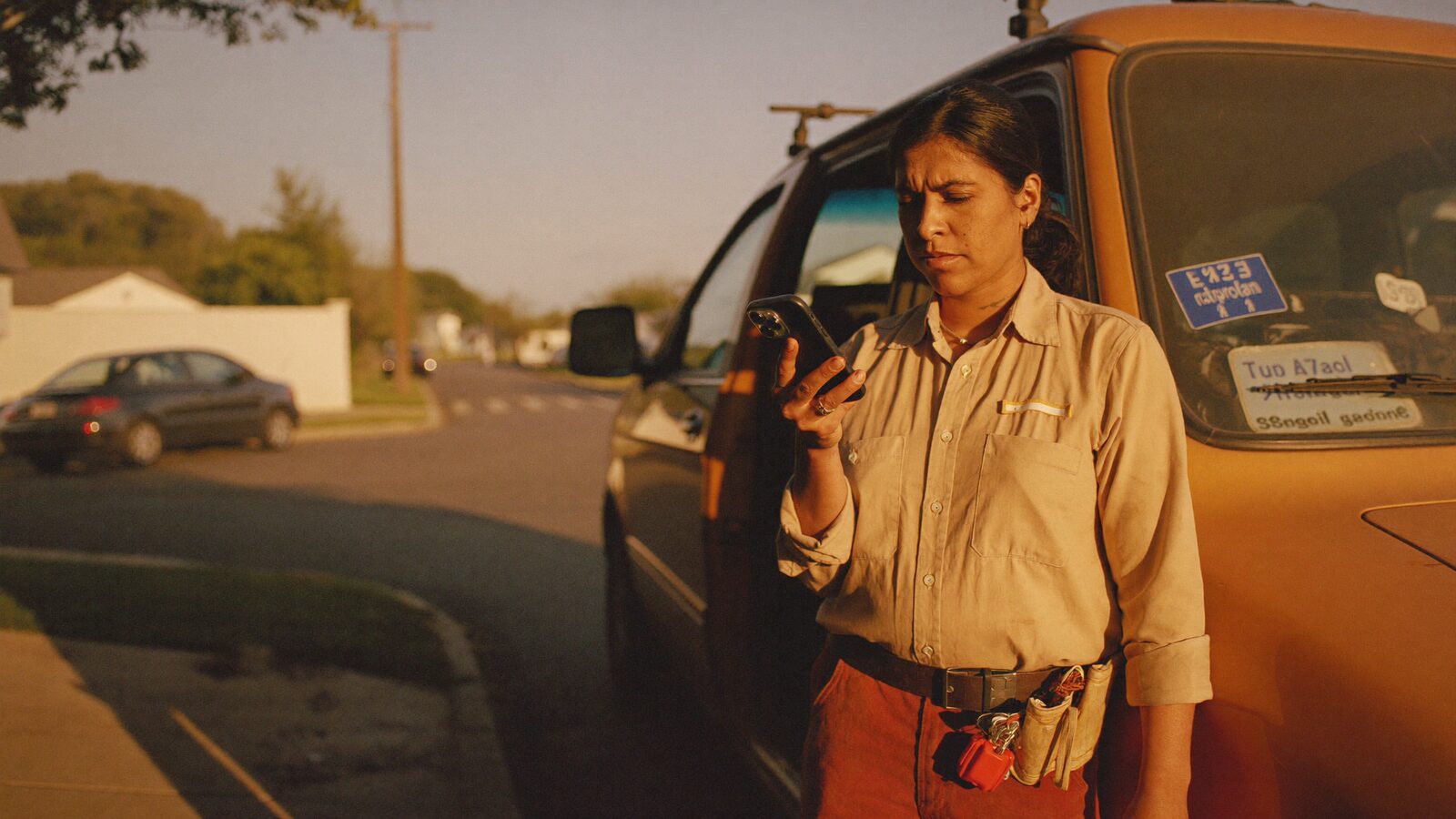 A South Asian woman electrician pauses beside her service van in late-afternoon light, glancing at a ringing phone