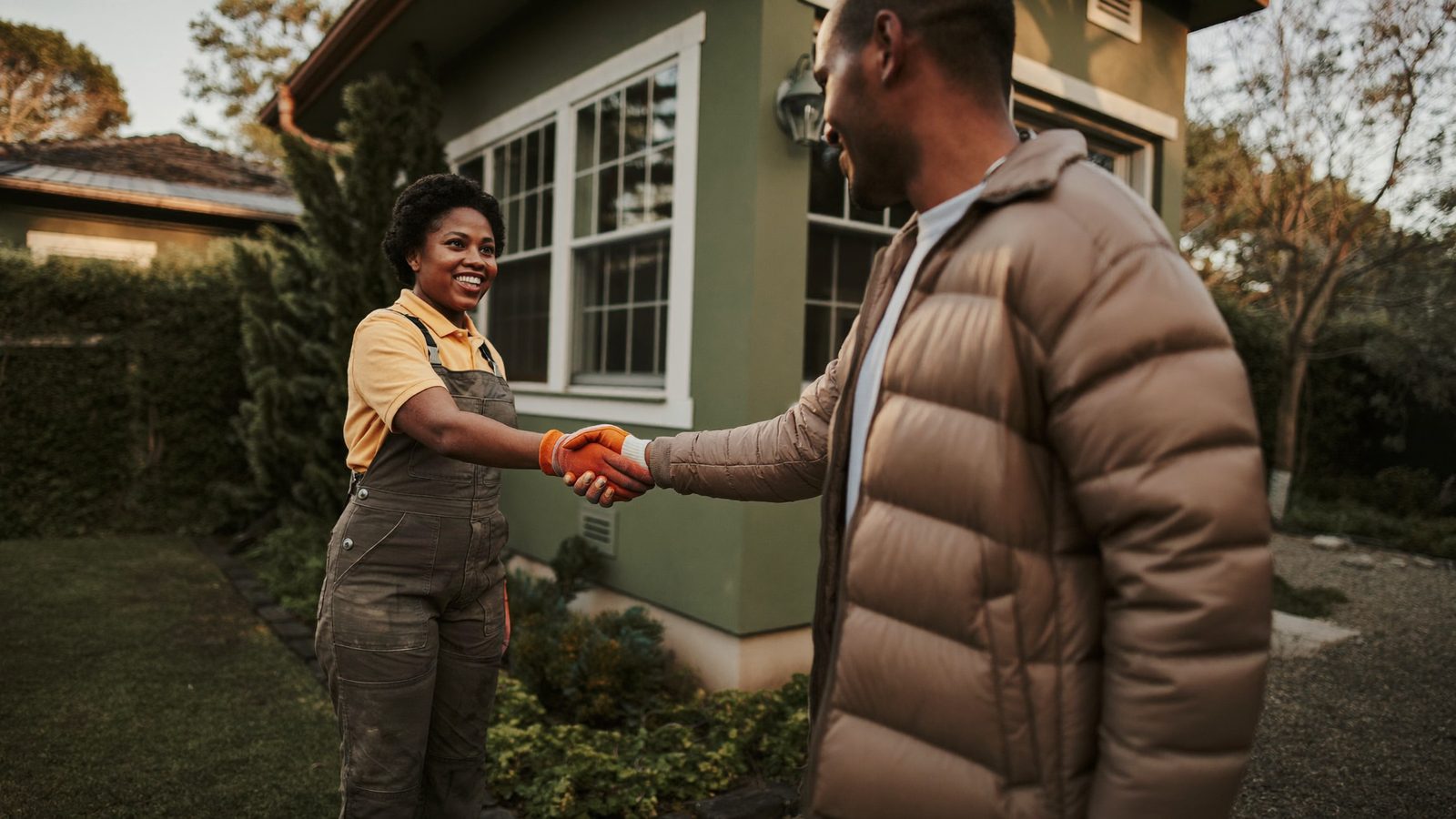 A Black woman landscaping business owner shakes hands with a smiling customer outside a freshly finished garden installation in warm afternoon light