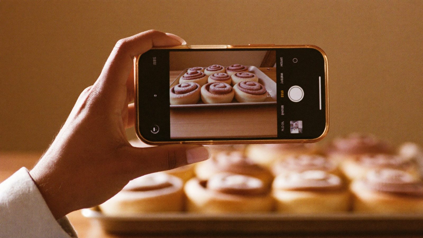 A hand holding a phone photographing a tray of fresh cinnamon rolls on a bakery counter for social media