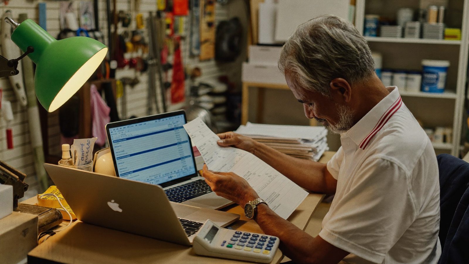 A hardware store owner reviewing invoices on a laptop at a cluttered back-office desk after closing