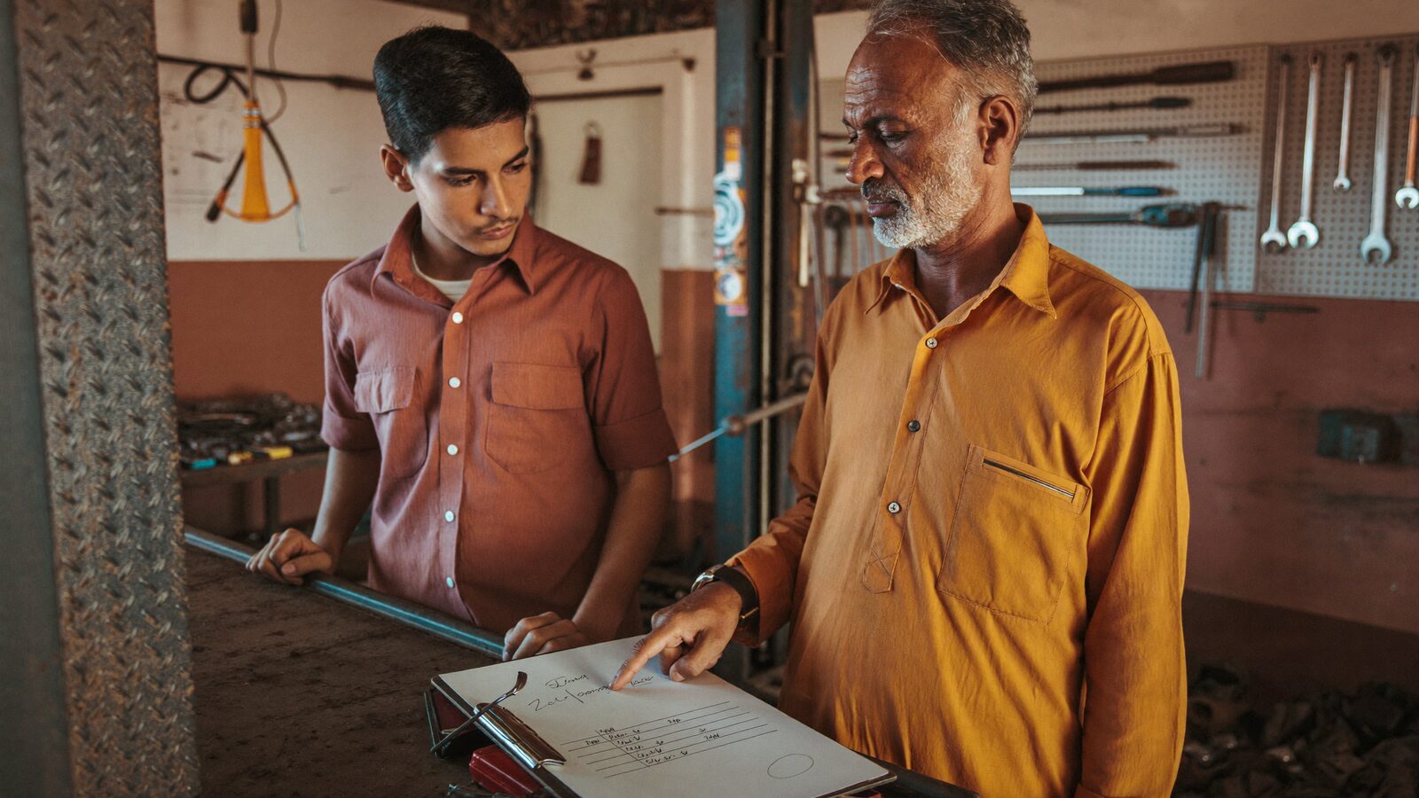 A small auto repair shop owner reviews a handwritten checklist with a younger apprentice in the garage bay