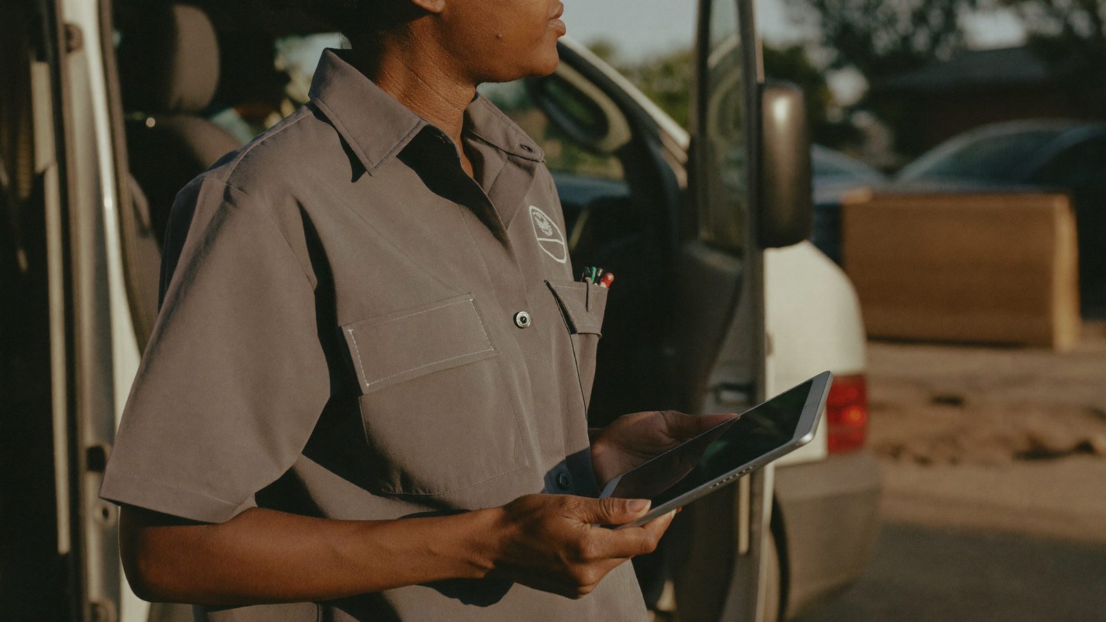 A tradeswoman in a plain uniform reviews a job estimate on a tablet beside her open service van
