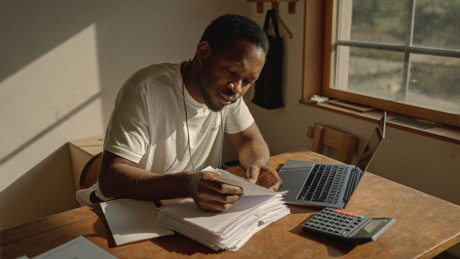 A small business owner reviewing paper invoices and a laptop at a cluttered desk in warm morning light